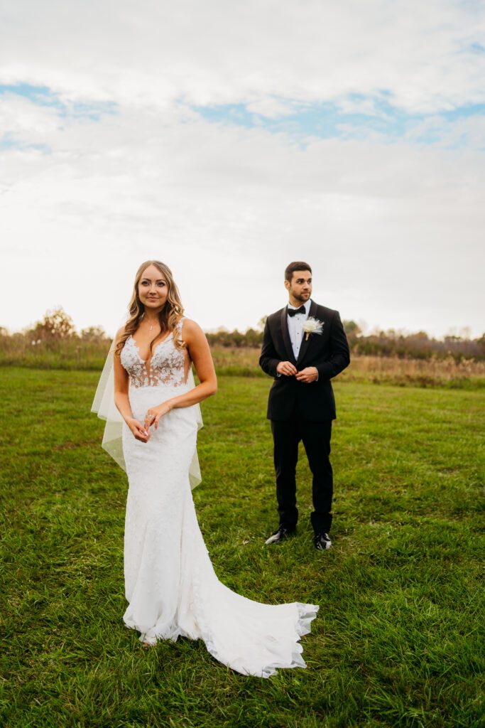 Bride in white dress standing in front of groom off to the side in a field