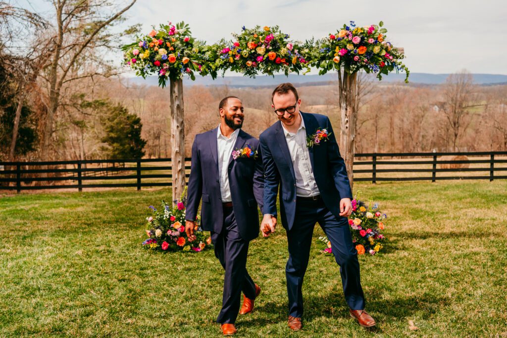 virginia-micro-wedding-lgbtq-couple-floral-arch.jpg Two grooms walk hand-in-hand under a floral arch in a scenic Virginia countryside micro wedding ceremony