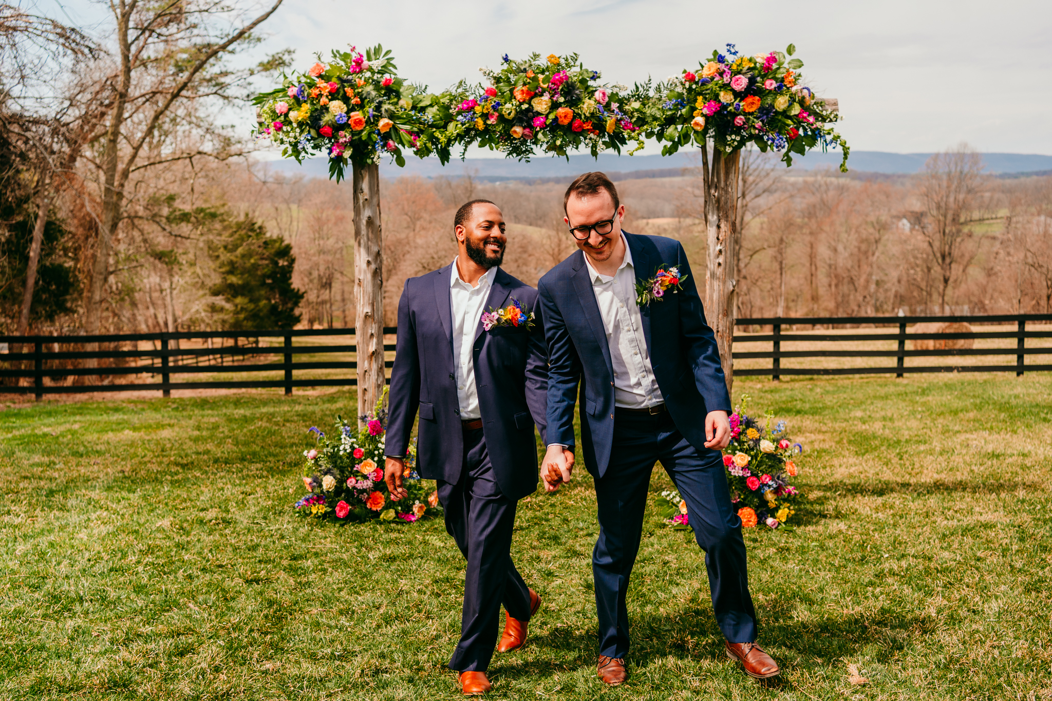 Two grooms walk hand-in-hand under a floral arch in a scenic Virginia countryside micro wedding ceremony