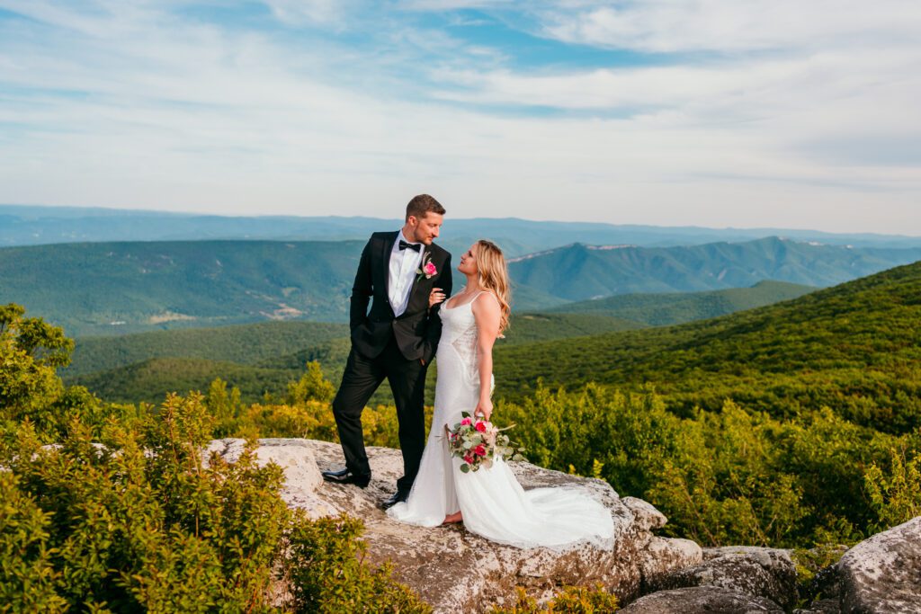Bride and groom smiling at each other on a rock ledge with lush green mountains stretching behind them