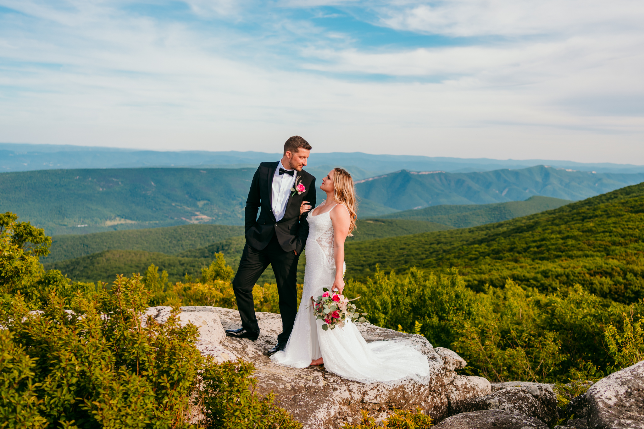 Bride and groom smiling at each other on a rock ledge with lush green mountains stretching behind them